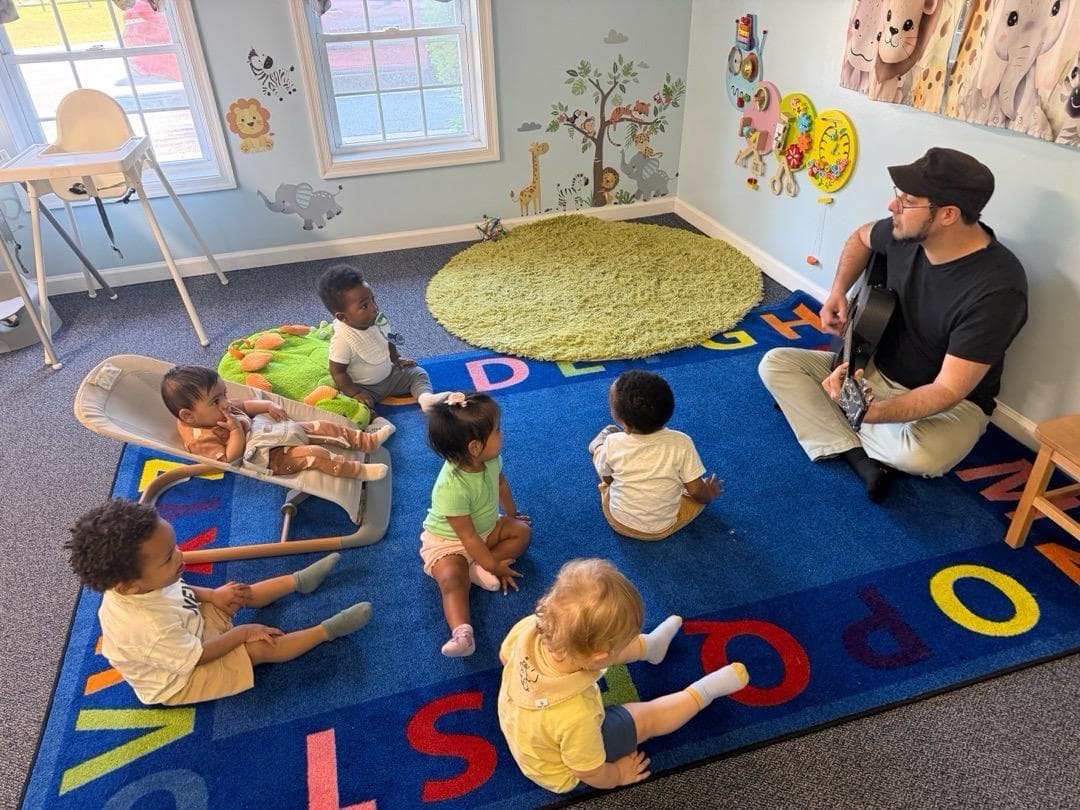 Infants sitting on an alphabet rug while a teacher plays guitar